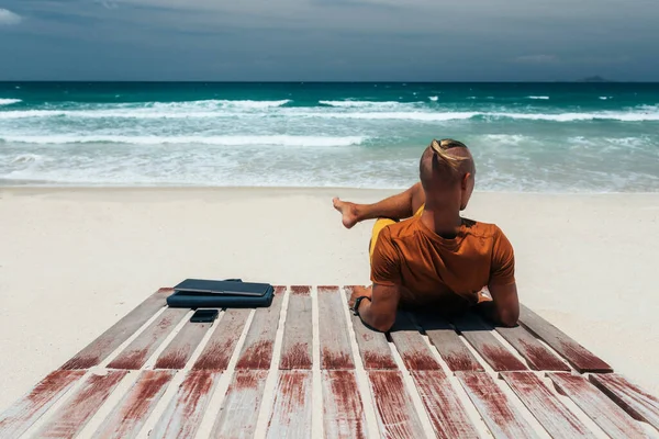 Young guy with long hair by the sea on a sunny day, working using his laptop, working remotely with a business. Bloner, freelancer, work through the Internet, work travel. Summer vacation. Sandy beach by the ocean with a wooden path. Copy place