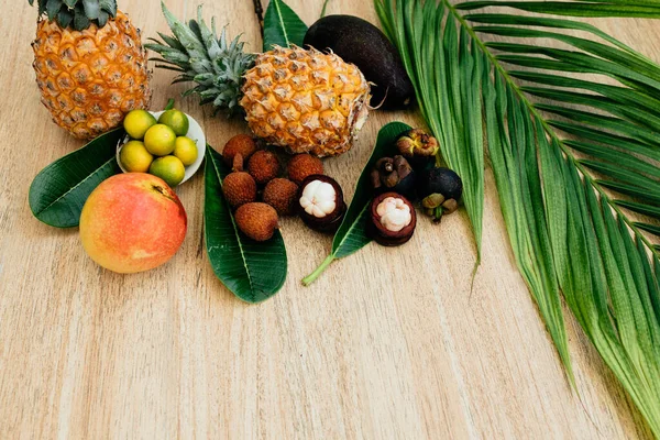 Düz bir zemin, üst manzara, kahverengi arka planda egzotik meyve ve palmiye yaprağı karışımı: ananas, Avokado, mango, mangosteen, lychee, citrus meyvesi. Sağlıklı bir diyet için harika bir set. Meyveli vejetaryenlik. Boşluğu kopyala, tahta arkaplan