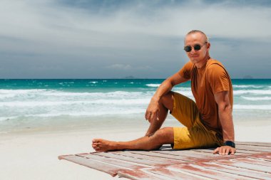 Genç adam yaz tatilinde tropik bir plajda oturur. Kameraya bakıyorum. Kumlu deniz manzarası, berrak bir yaz günü, beyaz bulutlu mavi bir gökyüzü. Yer kopyala