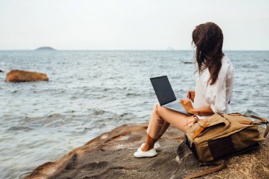 Genç başarılı kız dizüstü bilgisayar kullanarak çalışıyor, kayalık bir deniz kıyısında oturuyor. Seyahat ederken, blogcu, serbest çalışan, içerik planı hazırlayan, çevrimiçi çalışan bir iş kadını. Boşluğu kopyala