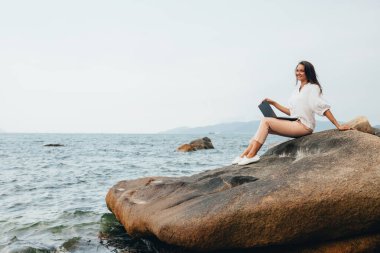 Genç, başarılı, gülümseyen bir kız dizüstü bilgisayar kullanıyor, kayalık bir deniz kıyısında oturuyor. Seyahat ederken, blog yazarken, internette çalışırken uzaktan çalışan bir iş kadını. Boşluğu kopyala