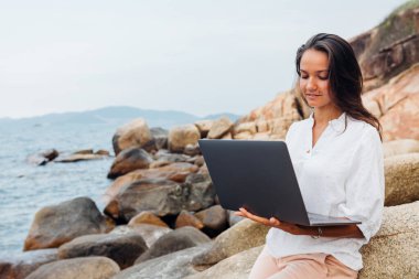 Genç, başarılı, gülümseyen bir kız dizüstü bilgisayar kullanıyor, kayalık bir deniz kıyısında oturuyor. Seyahat ederken, blogcu, serbest çalışan, içerik planı hazırlayan, çevrimiçi çalışan bir iş kadını. Boşluğu kopyala