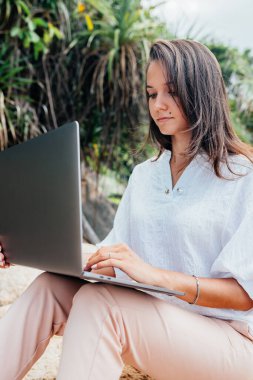Genç başarılı kız dizüstü bilgisayar kullanarak çalışıyor, kayalık bir deniz kıyısında oturuyor. Seyahat ederken, blogcu, serbest çalışan, içerik planı hazırlayan, çevrimiçi çalışan bir iş kadını. Boşluğu kopyala