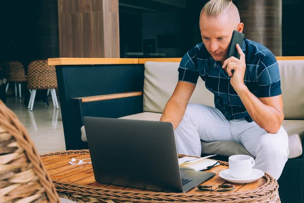 A young businessman, doing remote work while sitting in a cafe with a cup of coffee. Uses modern technology. Talking on the phone and looking at the laptop. Successful computer work on the Internet