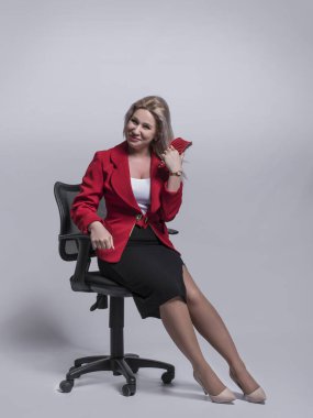 Güzel bir genç kız, sarışın, iş kadını kırmızı ceketli ve gri ve açık arkaplanda bir ofis sandalyesinde..