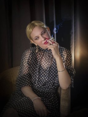 Genç, baştan çıkarıcı, duygusal, güzel bir sarışının, siyah, eski moda elbiseli bir kadının kavramsal portresi, bir otel odasının karanlık bir köşesinde, bir koltukta oturup sigara içiyor.