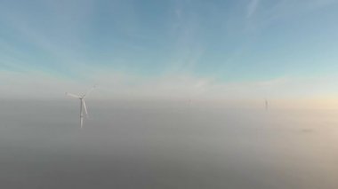 Rüzgar enerjisi çiftliği sis bulutlarının üzerinde, sabah, gün doğumunda. Dönen birkaç yel değirmeni, sisli hava. Yeşil, temiz, yenilenebilir enerji. Hava görüntüleri.