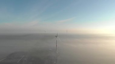 Güneşin doğuşunda sisli bir sabahta dönen üç yel değirmeni. Rüzgar enerjisi çiftliğinin inşaat alanı. Yeşil, temiz, yenilenebilir enerji. Hava görüntüleri.