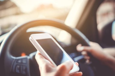 Direksiyondaki kadının eli akıllı telefon güneş ışığı arka planı kullanırken araba sürüyor..