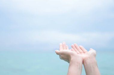 Kadın elleri, doğanın önünde dua eder gibi bir araya gelir. Temiz plaj ve mavi gökyüzü arka planı..