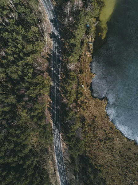 Drone footage flying over a road driving by towards a pine forest ,along the river in the ice on a sunny cold day.