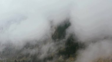 Drone flies through the autumn fog and clouds over the mountains towards the village and fjord.
