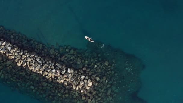 Drone shot of sea rocky stones. Man floats on a soup along a breakwater — ストック動画