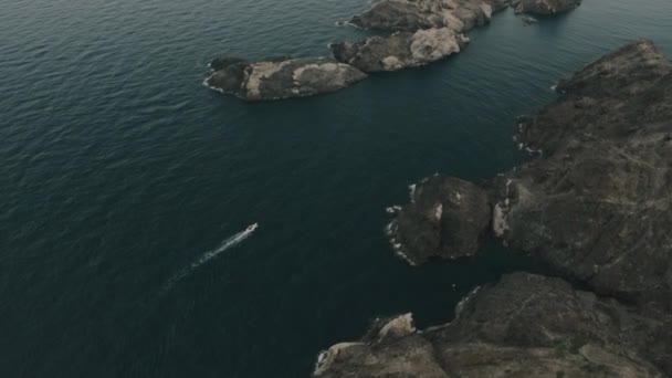 Drone flight over yacht in turquiose water of beautiful bay among rocks, small beach at mountain hill foot with lighthouse on top. — Stock Video