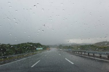 Araba ön camına yağmur damlası düşüyor, şehirde araba sürülüyor sağanak yağmur altında.
