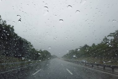 Araba ön camına yağmur damlası düşüyor, şehirde araba sürülüyor sağanak yağmur altında.