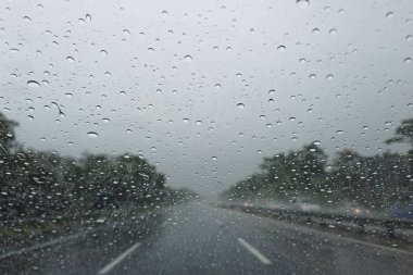 Araba ön camına yağmur damlası düşüyor, şehirde araba sürülüyor sağanak yağmur altında.