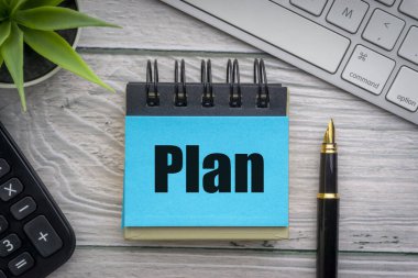 Dekoratif çiçek, hesap makinesi, dolmakalem, klavye ve tahta zemin üzerinde not defteri olan PLAN metni. İş ve iletişim kavramı