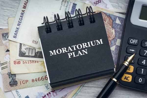 Dolma kalem, hesap makinesi, para birimi banknotları ve tahta zemin üzerinde not defteri olan MoratORIUM PLAN metni. İş ve ekonomi kavramı