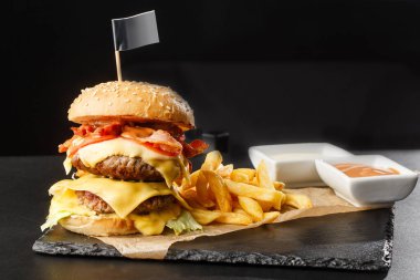 Siyah arka planda izole edilmiş patates kızartmalı büyük duble çizburger. Köfteli hamburger, pastırma, soğan, domates, marul turşusu, yıllanmış çedar, hardal, domates reçeli, mayonez. Yatay