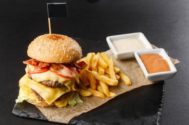 Siyah arka planda izole edilmiş patates kızartmalı büyük duble çizburger. Köfteli hamburger, pastırma, soğan, domates, marul turşusu, yıllanmış çedar, hardal, domates reçeli, mayonez. Yatay