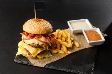 Siyah arka planda izole edilmiş patates kızartmalı büyük duble çizburger. Köfteli hamburger, pastırma, soğan, domates, marul turşusu, yıllanmış çedar, hardal, domates reçeli, mayonez. Yatay