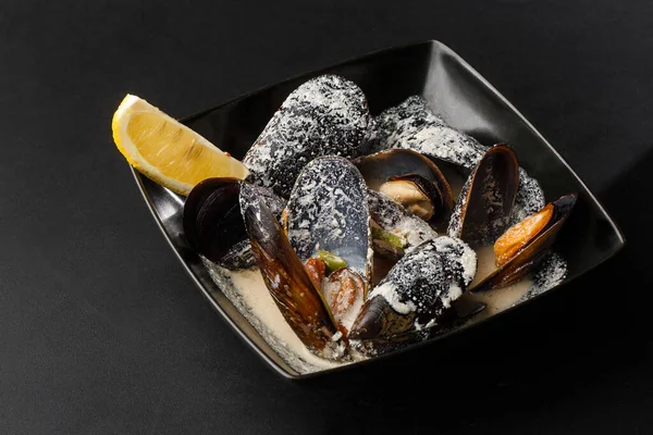 Vers gekookte schaal- en schelpdieren mosselen met room, wijn, kaassaus en citroen in een zwarte kom, close-up, studio shot. Een klassiek gerecht in België, Frankrijk en Nederland. Restaurant eten. Horizontaal — Stockfoto