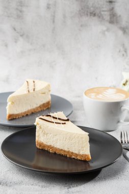 Cheesecake New York ya da beyaz masada bir fincan kahveli klasik cheesecake. Yakından bak. Lezzetli bir kahvaltı. Siyah tabakta bir parça kek, beyaz mermer arka planda beyaz bardak. Dikey fotoğraf.
