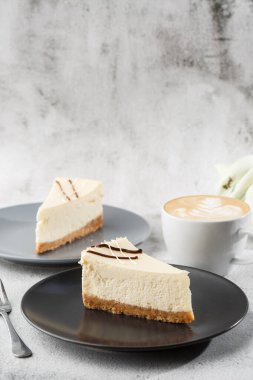 Cheesecake New York ya da beyaz masada bir fincan kahveli klasik cheesecake. Yakından bak. Lezzetli bir kahvaltı. Siyah tabakta bir parça kek, beyaz mermer arka planda beyaz bardak. Dikey fotoğraf.