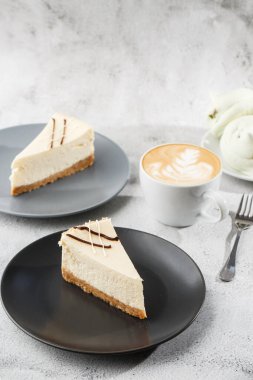 Cheesecake New York ya da beyaz masada bir fincan kahveli klasik cheesecake. Yakından bak. Lezzetli bir kahvaltı. Siyah tabakta bir parça kek, beyaz mermer arka planda beyaz bardak. Dikey fotoğraf.