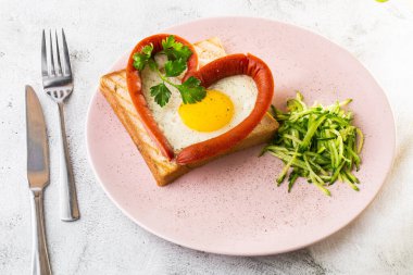 Kalp şeklinde çırpılmış yumurtalar, beyaz bir tabakta sosisli, ekşi ekmek, beyaz mermer arka planda izole edilmiş. Ev yapımı yemek. Lezzetli bir kahvaltı. Seçici odaklanma. Hotizontal fotoğraf.