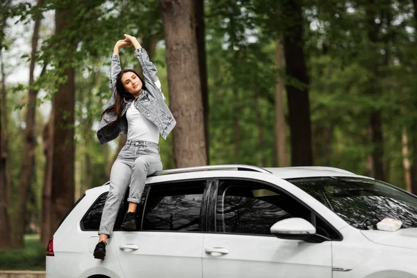 Güzel, seksi moda esmer kadın modelinin portresi kot ceketli, beyaz lüks bir arabanın üzerinde oturuyor ve rahatlıyor. Moda konsepti. Arabalı kız fotoşopu fikri. Yatay.