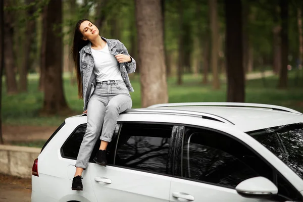 Portret pięknej seksownej brunetki modela model w dżinsach kurtka siedzi na luksusowym białym samochodzie i relaksuje. Koncepcja mody. Pomysł na sesję zdjęciową z samochodem. Poziomy. — Zdjęcie stockowe