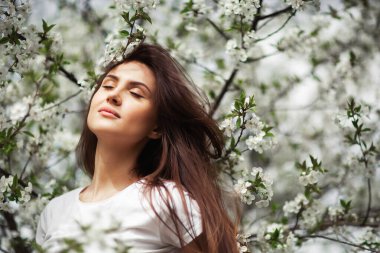 Doğal parktaki çiçek açan elma ağacının altında duran kız. Moda konsepti. Kot elbiseli zarif genç bayan güneşli bahar gününde çiçek açan bahçenin tadını çıkarıyor. Doğal güzellik makyajı. Yatay