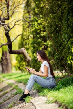 Çekici kız hafta sonu doğada dinleniyor. Parkta gölün kenarında oturan kot elbiseli kız. Kafkas esmer, genç bir kadın tek başına akıllı telefondan mesaj yolluyor. Teknoloji ve cihazlar.