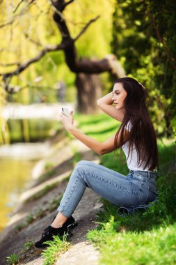 Çekici kız hafta sonu doğada dinleniyor. Parkta gölün kenarında oturan kot elbiseli kız. Kafkas esmer, genç bir kadın dışarıda akıllı telefondan selfie çekiyor. Teknoloji ve aletler. Ver.