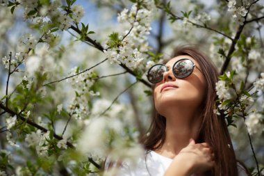 Doğal parktaki çiçek açan elma ağacının altında duran kız. Moda konsepti. Kotlu ve güneş gözlüklü zarif genç bayan güneşli bahar gününde çiçek açan bahçenin tadını çıkarıyor. Doğal güzellik makyajı. Hori.