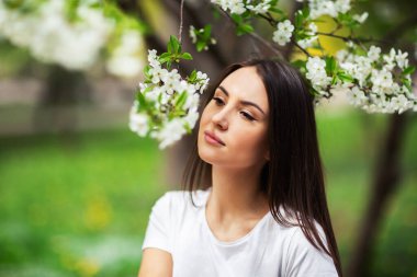 Doğal parktaki çiçek açan elma ağacının altında duran kız. Moda konsepti. Kot elbiseli zarif genç bayan güneşli bahar gününde çiçek açan bahçenin tadını çıkarıyor. Doğal güzellik makyajı. Yatay
