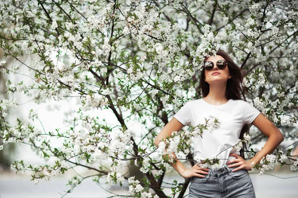 Menina de pé sob a árvore de maçã florescente no parque natural. Conceito de moda. Menina elegante em jeans roupas e óculos de sol desfrutando de jardim florido no dia ensolarado.Maquiagem de beleza natural. Hori... — Fotografia de Stock
