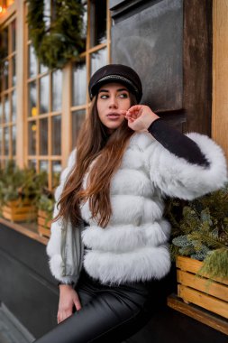 Doğal kısa beyaz kürk giyen güzel beyaz kadın Noel vitrininin yanındaki bir portre için poz veriyor. Noel dekoru. Moda. Tarz. Doğal kürkten yapılmış kışlık giysiler. Dikey
