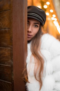 Doğal kısa beyaz kürk giyen güzel beyaz kadın Noel vitrininin yanındaki bir portre için poz veriyor. Noel dekoru. Moda. Tarz. Doğal kürkten yapılmış kışlık giysiler. Dikey