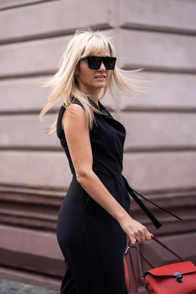 Открытая мода. Блондинка в модном стильном черном комбинезоне, солнечных очках и красной сумке осенний образ жизни на фоне размытых желто-зеленых деревьев в парке. Вертикаль — стоковое фото