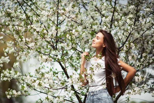 Fille debout sous le pommier en fleurs dans le parc naturel. Concept de mode. Elégante jeune femme en jeans vêtements appréciant jardin fleuri au jour ensoleillé du printemps.Maquillage beauté naturelle. Horizontal — Photo