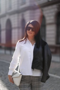 Sonbahar modası açık havada. Modaya uygun beyaz gömlekli, pantolonlu, ceketli, güneş gözlüklü parktaki bulanık sarı-yeşil ağaçların arka planında sonbahar yaşam tarzı olan kız. Dikey