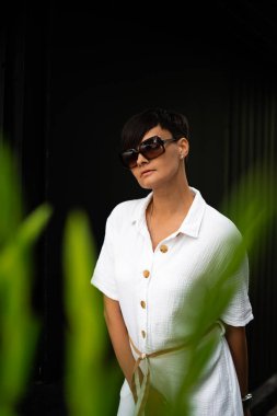 Kısa esmer saçlı, beyaz elbiseli ve güneş gözlüklü güzel bir kadın. Moda sokak fotoğrafçılığı. Koyu arkaplanda poz veren manken.