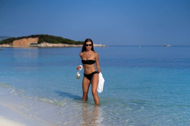 Alkolle poz veren genç bir kız. Atletik, mutlu, modaya uygun, seksi siyah yüzme kıyafetleriyle koşan, güneşin tadını çıkaran bir kadın. Sağlıklı bir yaşam tarzı. Sahilde eğlenceli bir yürüyüş. Mükemmel fitness vücut şekilleri.