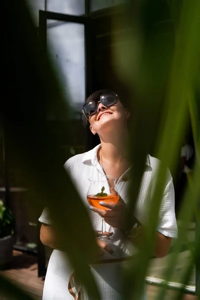 Belle femme aux cheveux bruns courts en vêtements blancs et lunettes de soleil. Photographie de rue de mode. Modèle de mode posant avec cocktail aperol sur fond bois . — Photo