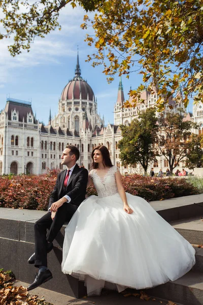 Nevěsta a ženich se objímají na staré ulici. Svatební dvojice sedí v Budapešti poblíž parlamentu. Kavkazský šťastný romantický mladý pár oslavující své manželství. Svatební a milostný koncept. — Stock fotografie