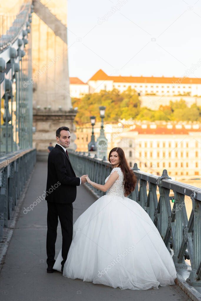 Bride and groom hugging in the old town street. Wedding couple walking on Szechenyi Chain Bridge, Hungary. Happy romantic young couple celebrating their marriage. Wedding and love concept.
