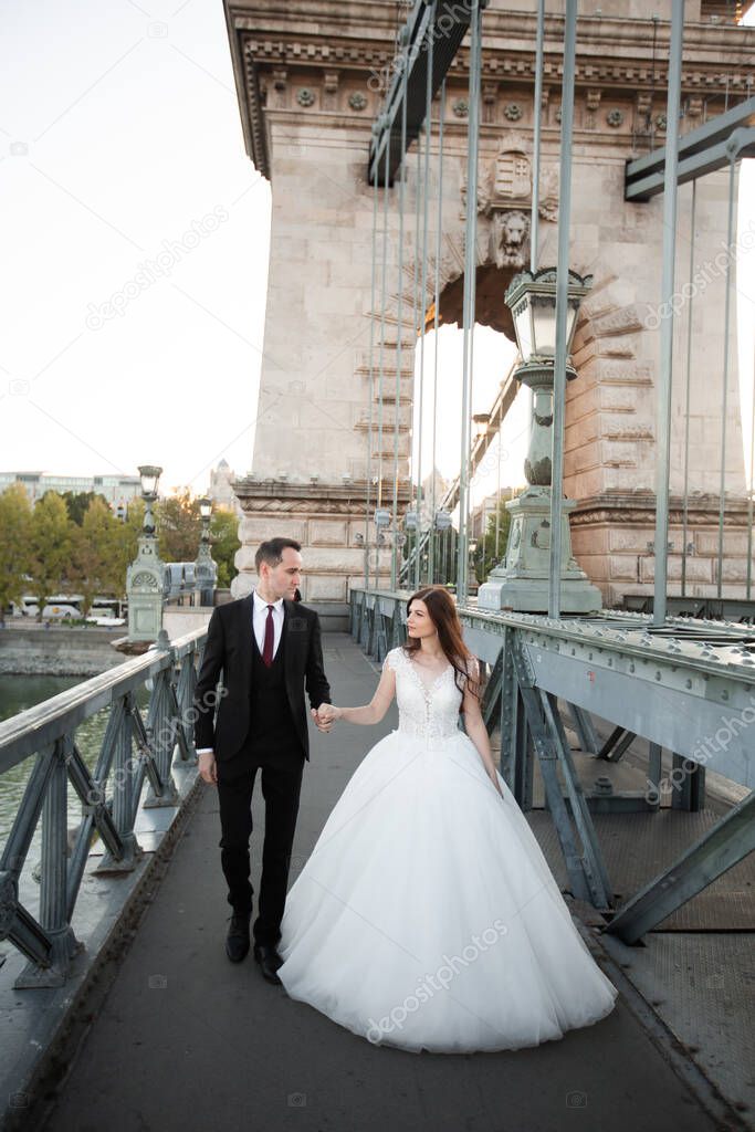 Bride and groom hugging in the old town street. Wedding couple walking on Szechenyi Chain Bridge, Hungary. Happy romantic young couple celebrating their marriage. Wedding and love concept.